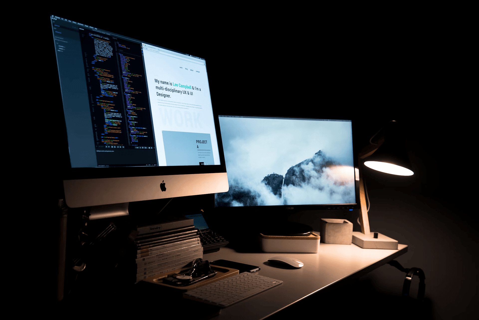 A stylish desk setup featuring two monitors displaying code and a web page, a desk lamp, and organized accessories in a dark setting.