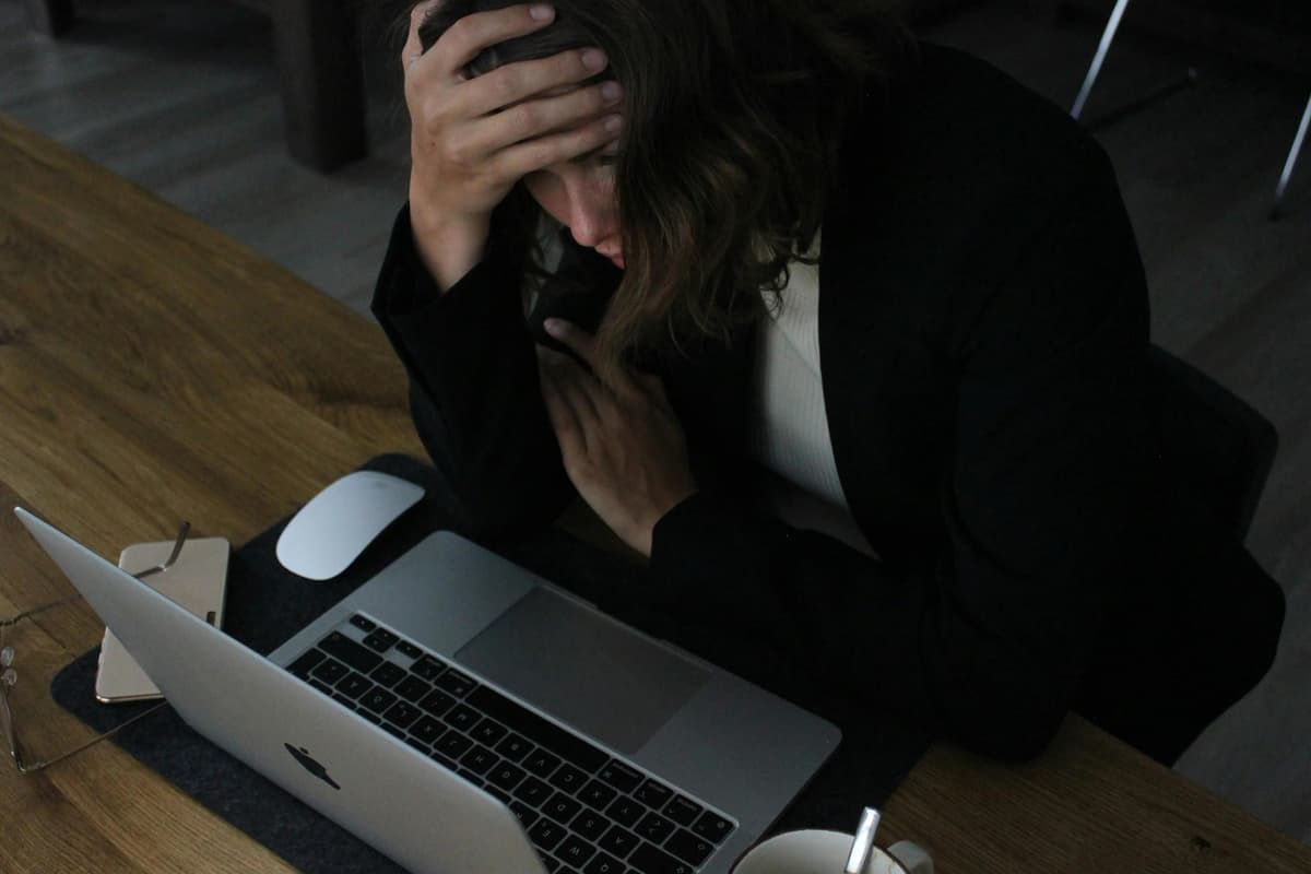 Stressed woman in business attire sitting at a wooden desk with her hand on her forehead, looking down at an open laptop. A smartphone, computer mouse, glasses, and a coffee cup are also on the desk.