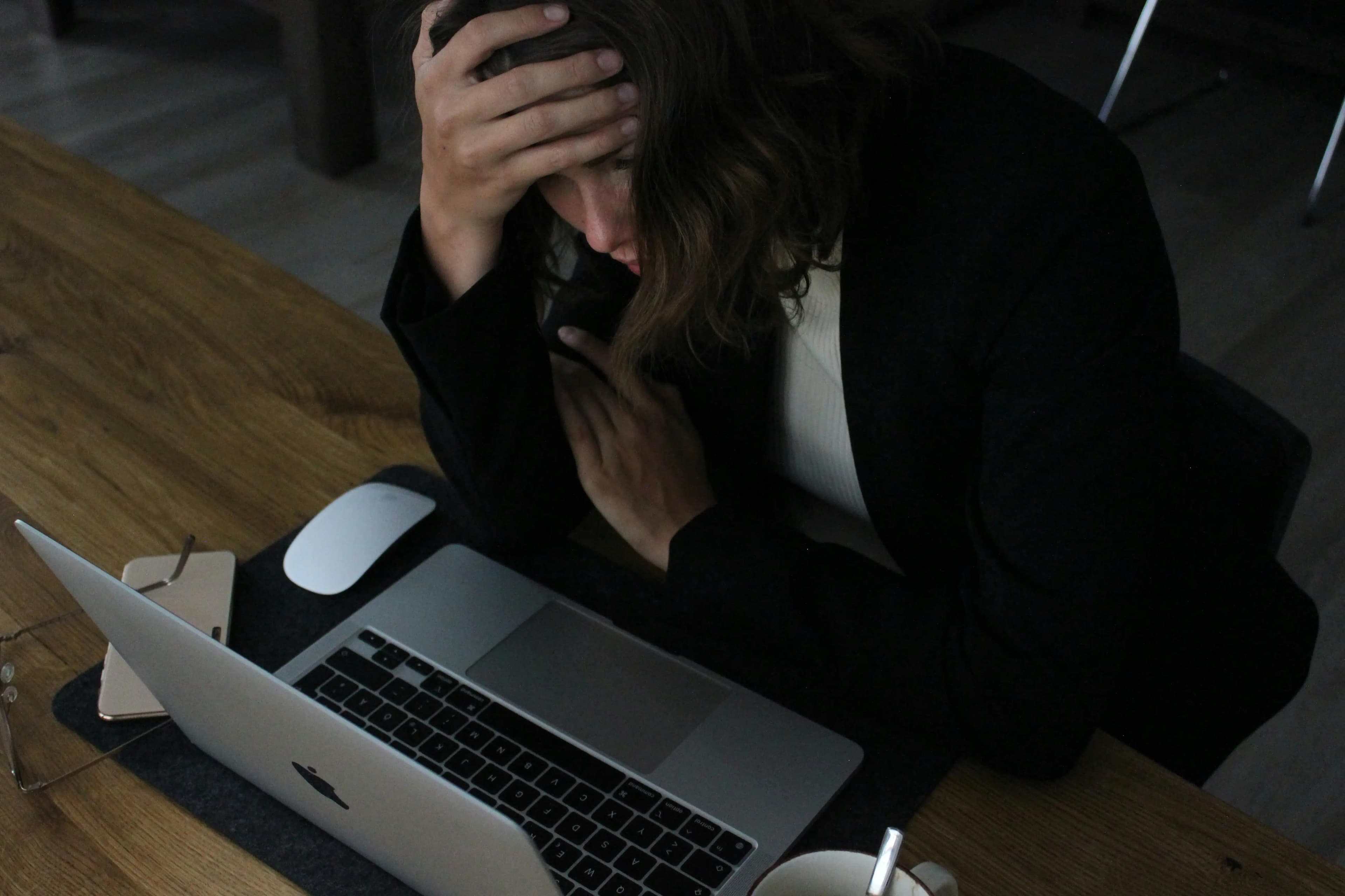 Stressed woman in business attire sitting at a wooden desk with her hand on her forehead, looking down at an open laptop. A smartphone, computer mouse, glasses, and a coffee cup are also on the desk.
