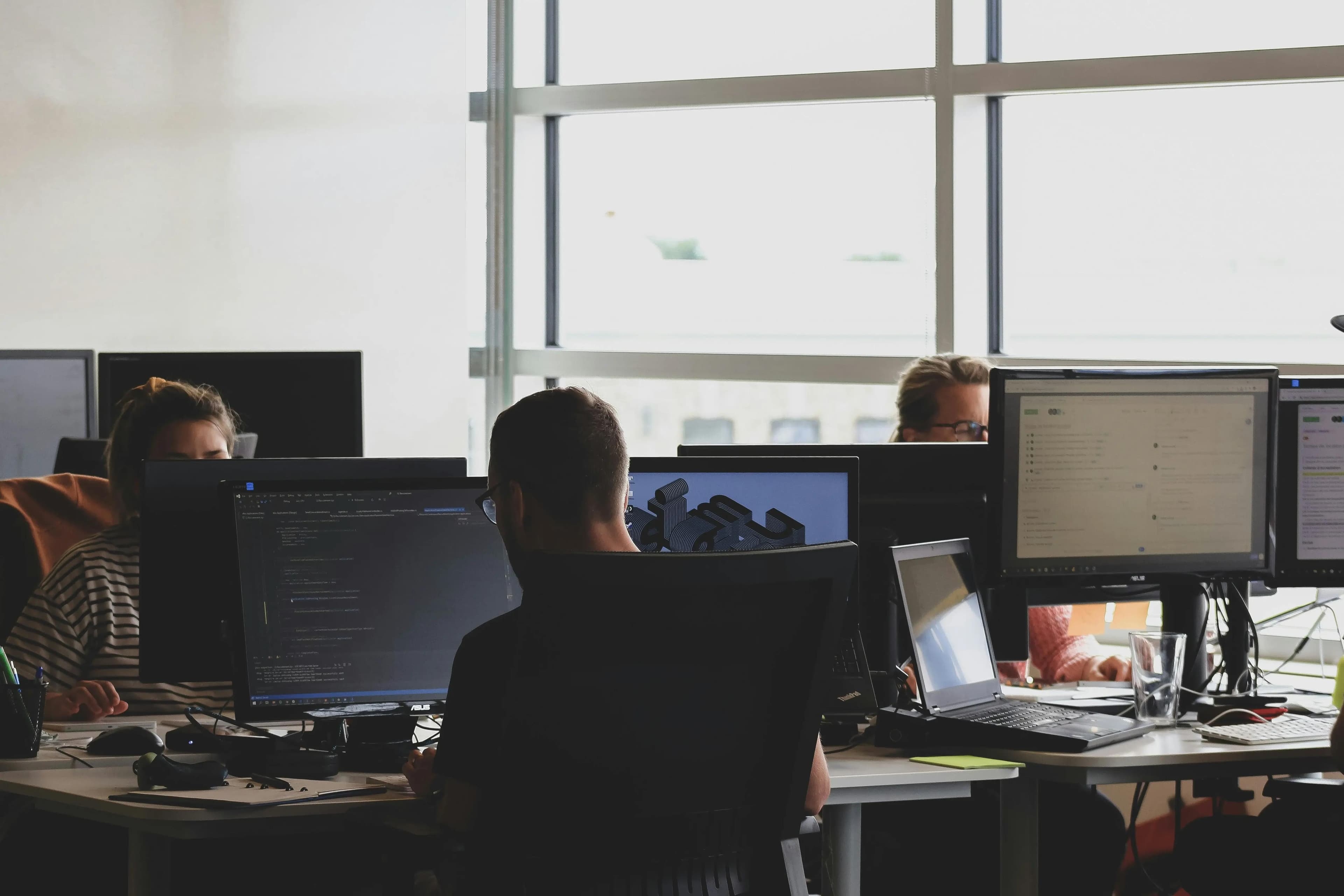 People working at desks in a modern office, each focused on multiple computer monitors displaying code, 3D models, and data interfaces.