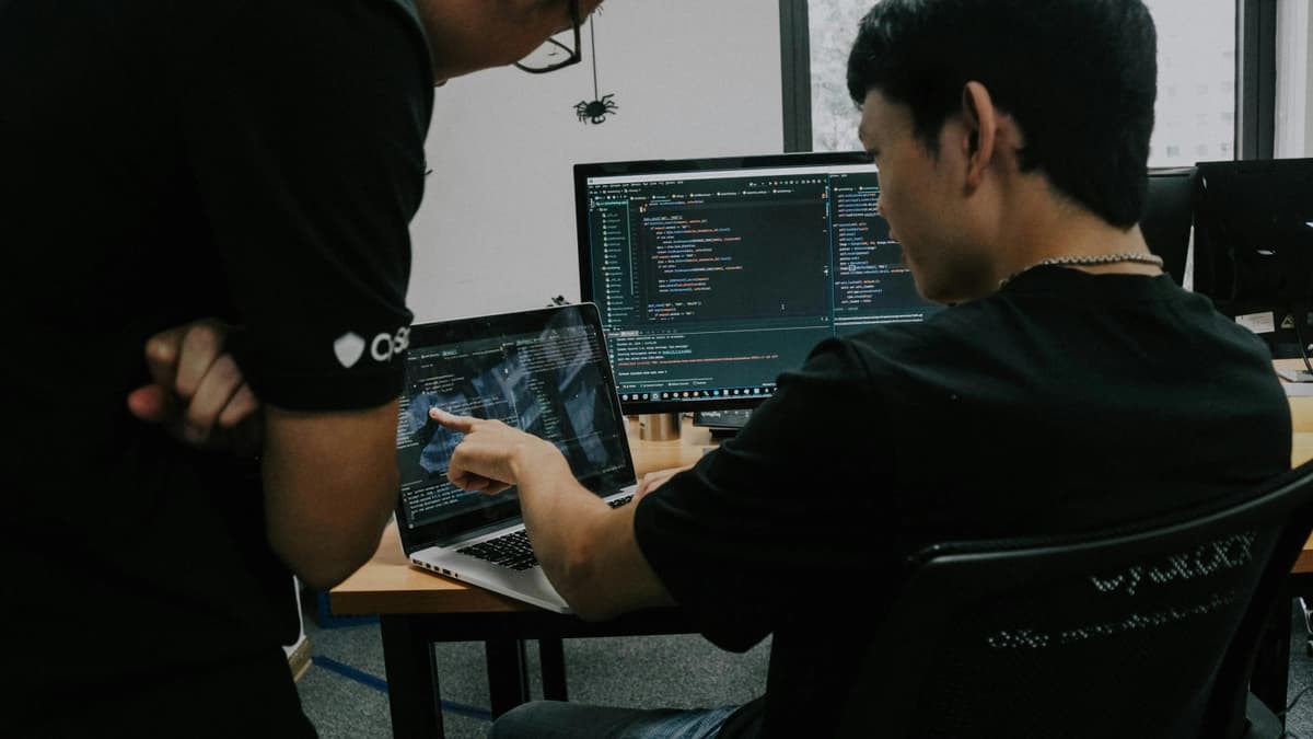Two developers collaborating in an office, one pointing at code on a laptop screen while the other views code on a desktop monitor.
