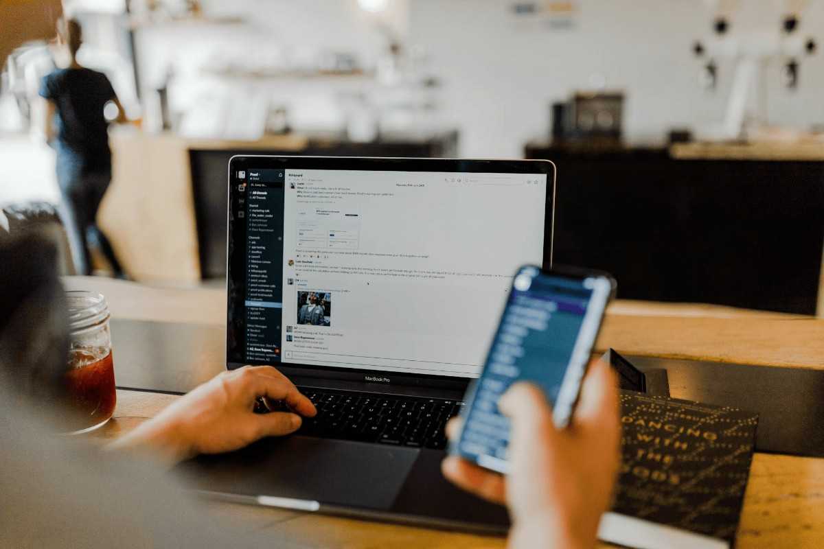 A person working on a laptop at a café, typing on a messaging app while holding a smartphone displaying a notification.